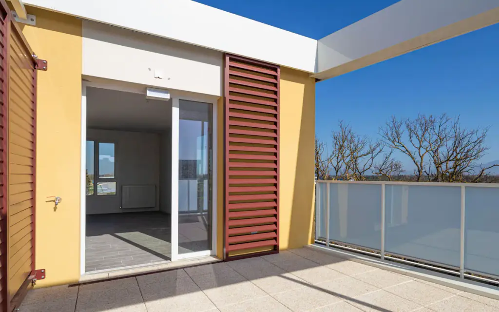 Terrasse d’un logement neuf : volet coulissant à lames rouge brique ouvert le long d’une façade jaune, porte-fenêtre ouverte sur une pièce vide, garde-corps en verre dépoli, ciel bleu.