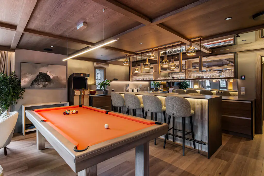 Billard Maison Toulet en chêne fumé à drap orange, billes en place, dans une salle au plafond de bois devant un bar à tabourets.