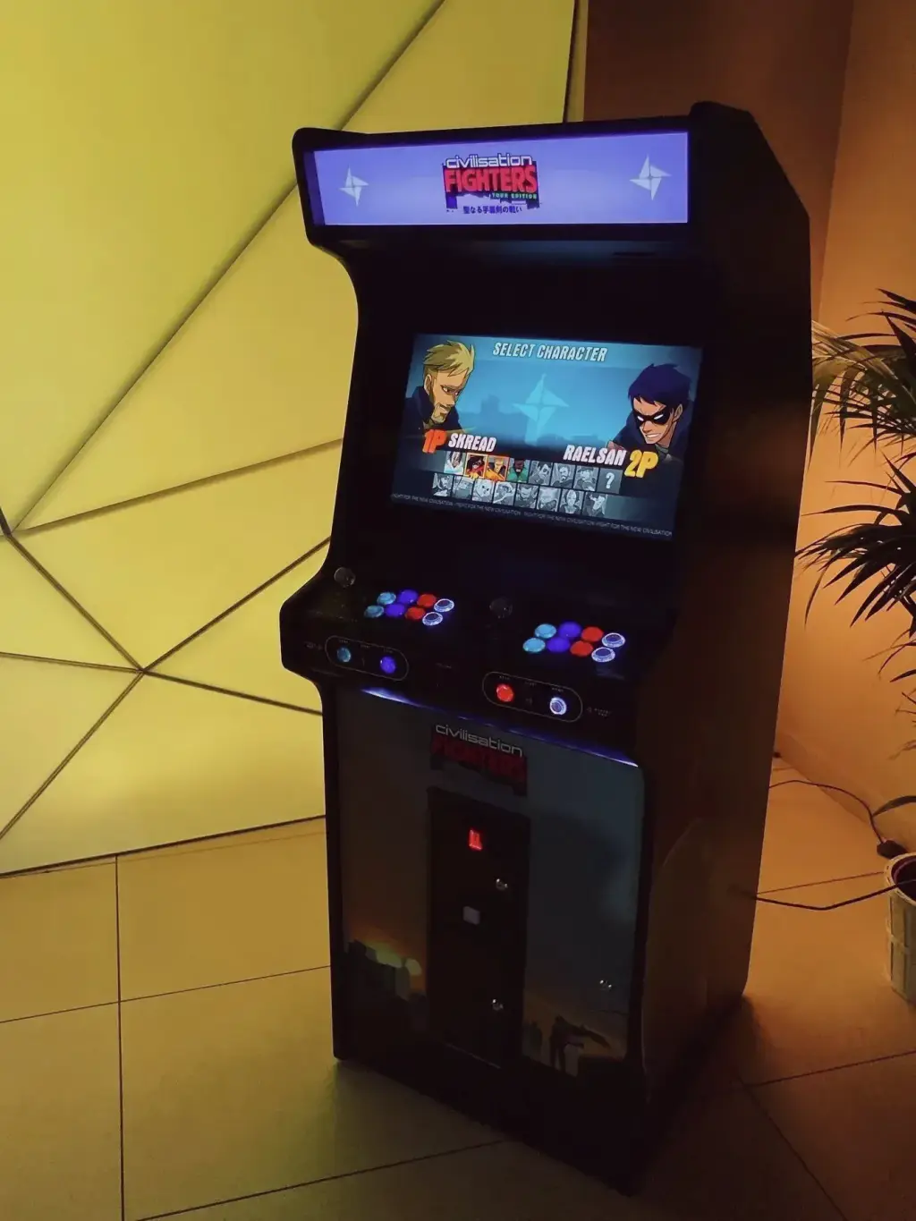 Une borne d’arcade Neo Legend noire, debout, vue de trois quarts : fronton éclairé, écran allumé, pupitre à deux manches aux boutons lumineux, devant un mur éclairé de jaune ; une plante dans l’ombre.