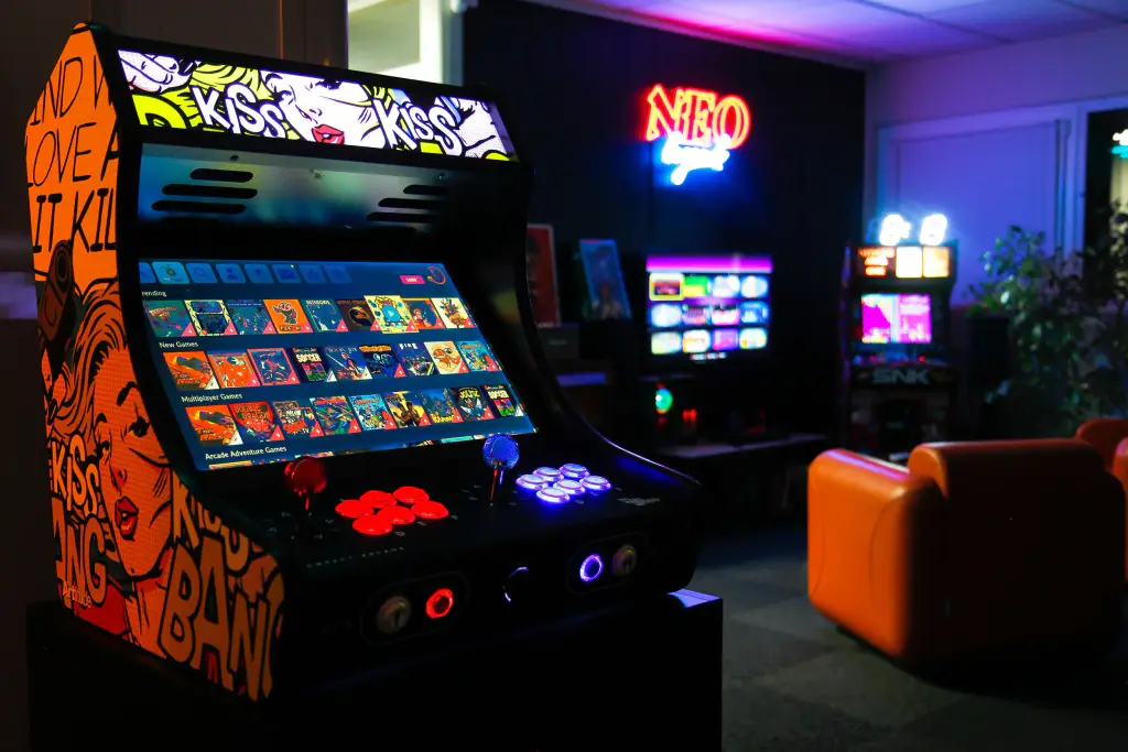 Une borne d’arcade Neo Legend à décor pop art orange, écran allumé sur la liste des jeux, boutons rouges et bleus éclairés ; au fond, une salle sombre avec d’autres bornes, une enseigne au néon et des fauteuils orange.