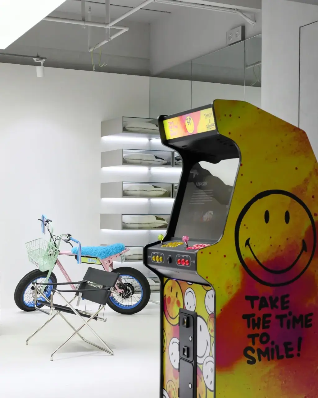 Une borne d’arcade Neo Legend au décor Smiley jaune et rose, de profil, dans un loft blanc : étagères éclairées, une chaise pliante et un vélo au fond.
