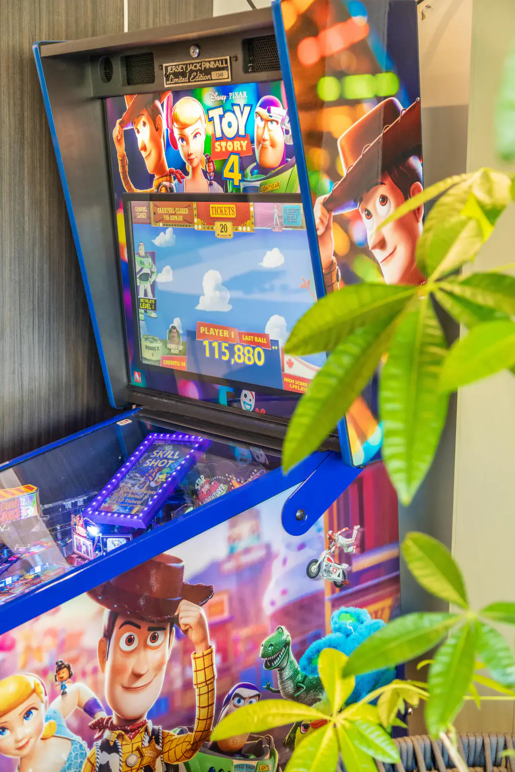 Fronton du flipper Toy Story 4 de Jersey Jack Pinball : plaque « Limited Edition », écran allumé affichant un score de 115 880 points, haut du plateau sous la vitre et flanc illustré de la caisse, derrière le feuillage d’une plante.