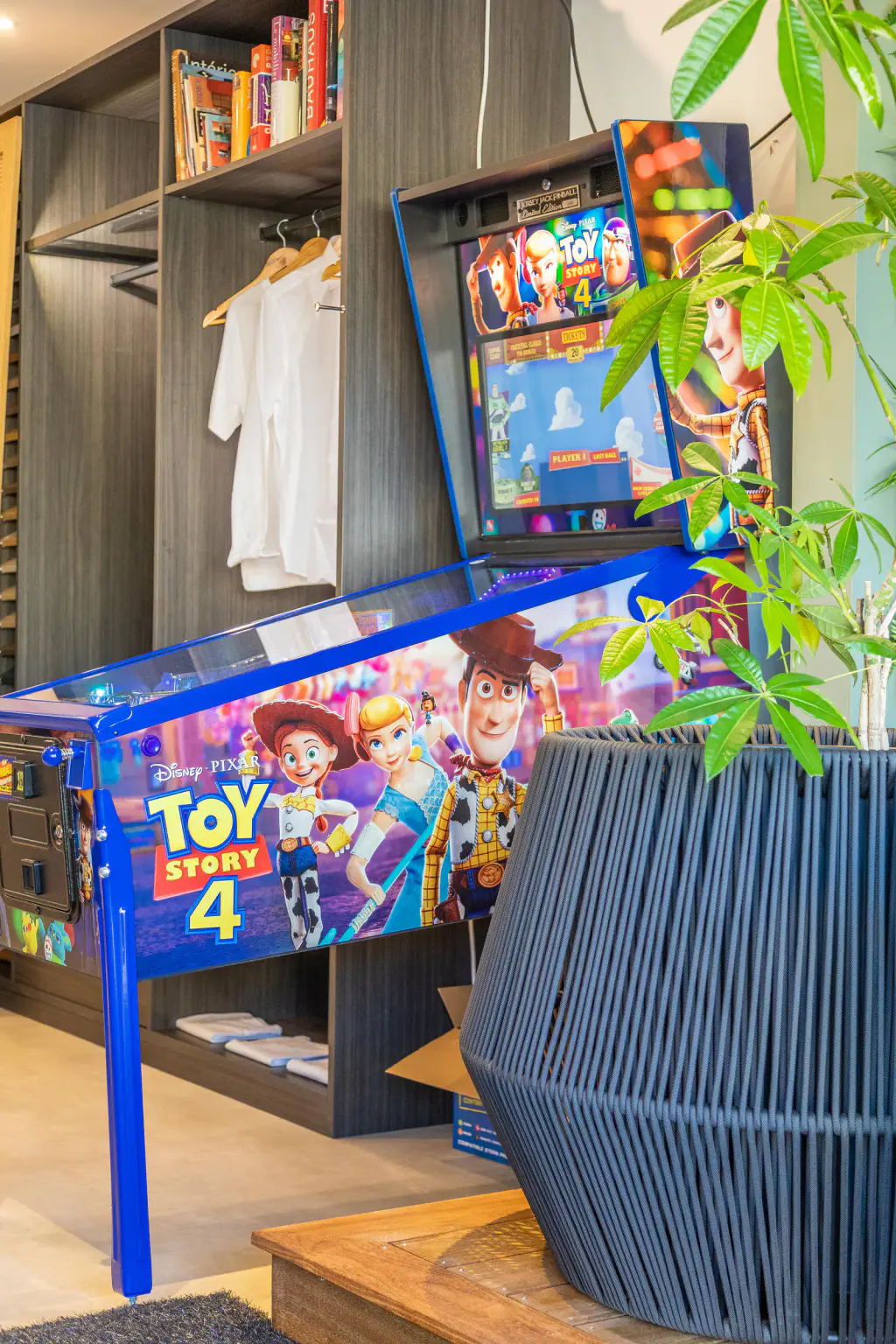 Le flipper Toy Story 4 de Jersey Jack Pinball au showroom LumiDesign de Porticcio, vu de trois quarts devant les rangements en bois gris, une plante verte au premier plan.