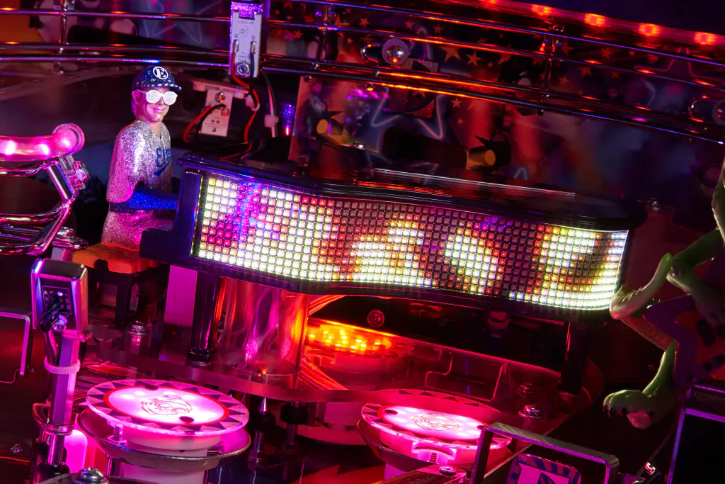 Le plateau allumé d’un flipper Jersey Jack Pinball Elton John : la figurine du chanteur à son piano, une enseigne de lumières, des bumpers rose et rouge dans la pénombre.