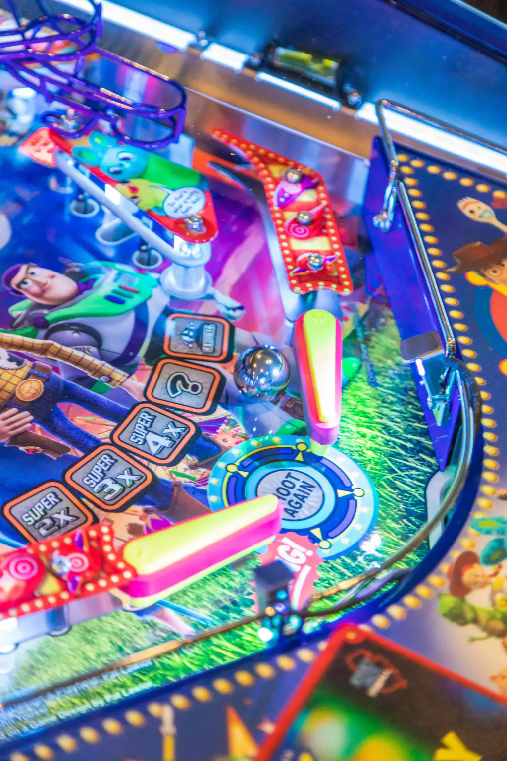 Bas du plateau du flipper Toy Story 4 : une bille d’acier au-dessus des deux batteurs jaunes et roses, l’insert « Shoot again », les inserts « Super 2x », « 3x » et « 4x » et le couloir de sortie de droite.