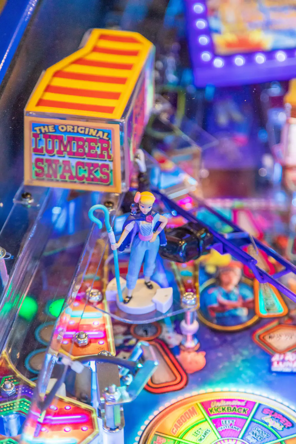 Détail du plateau : une figurine de bergère tenant sa houlette bleue, sous un décor en forme de camion rayé jaune et rouge marqué « Lumber Snacks », la roue multicolore floue au premier plan.