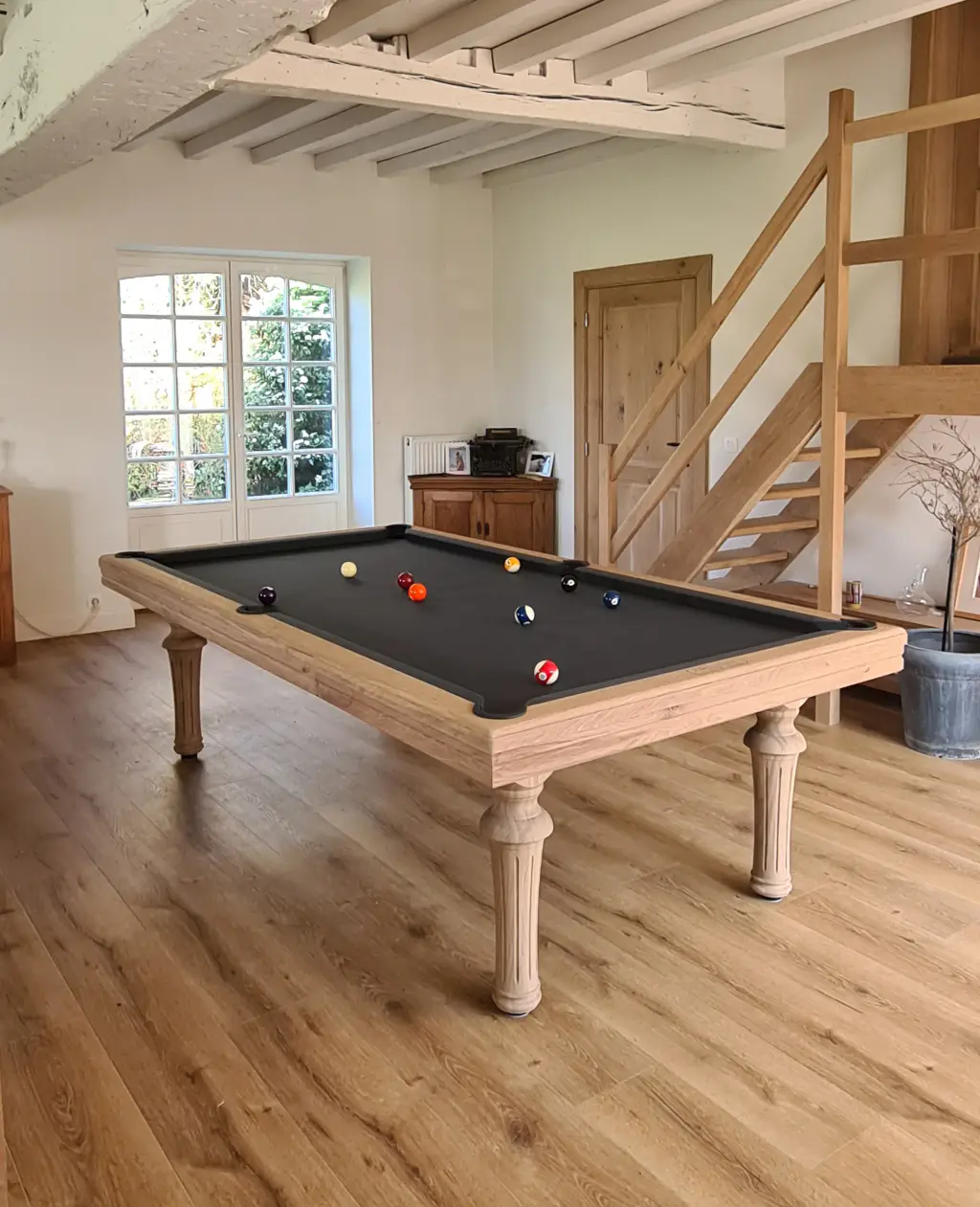 Un billard Maison Toulet en chêne clair aux pieds tournés et cannelés, drap noir et billes en jeu, au milieu d’une pièce aux poutres blanchies : parquet de chêne, porte-fenêtre sur le jardin, escalier de bois et vieux buffet.