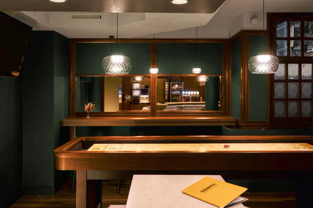 Un long shuffleboard Maison Toulet en bois verni, palets posés, dans une salle de bar aux boiseries vert sombre : suspensions de verre, comptoir et miroirs au fond.