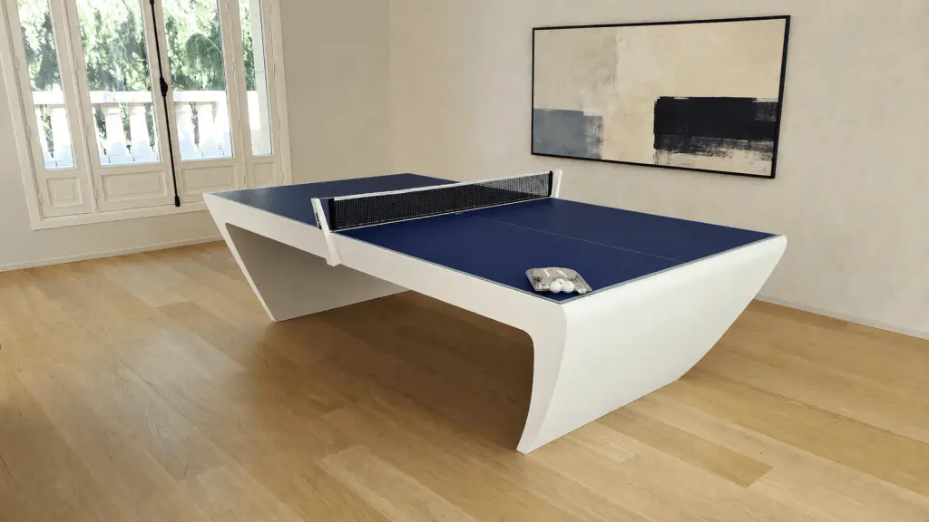 Une table de ping-pong Maison Toulet aux flancs blancs galbés et au plateau bleu, filet tendu, raquettes posées, au milieu d’une pièce vide au parquet clair : grande toile abstraite au mur, portes-fenêtres sur un balcon à balustres.