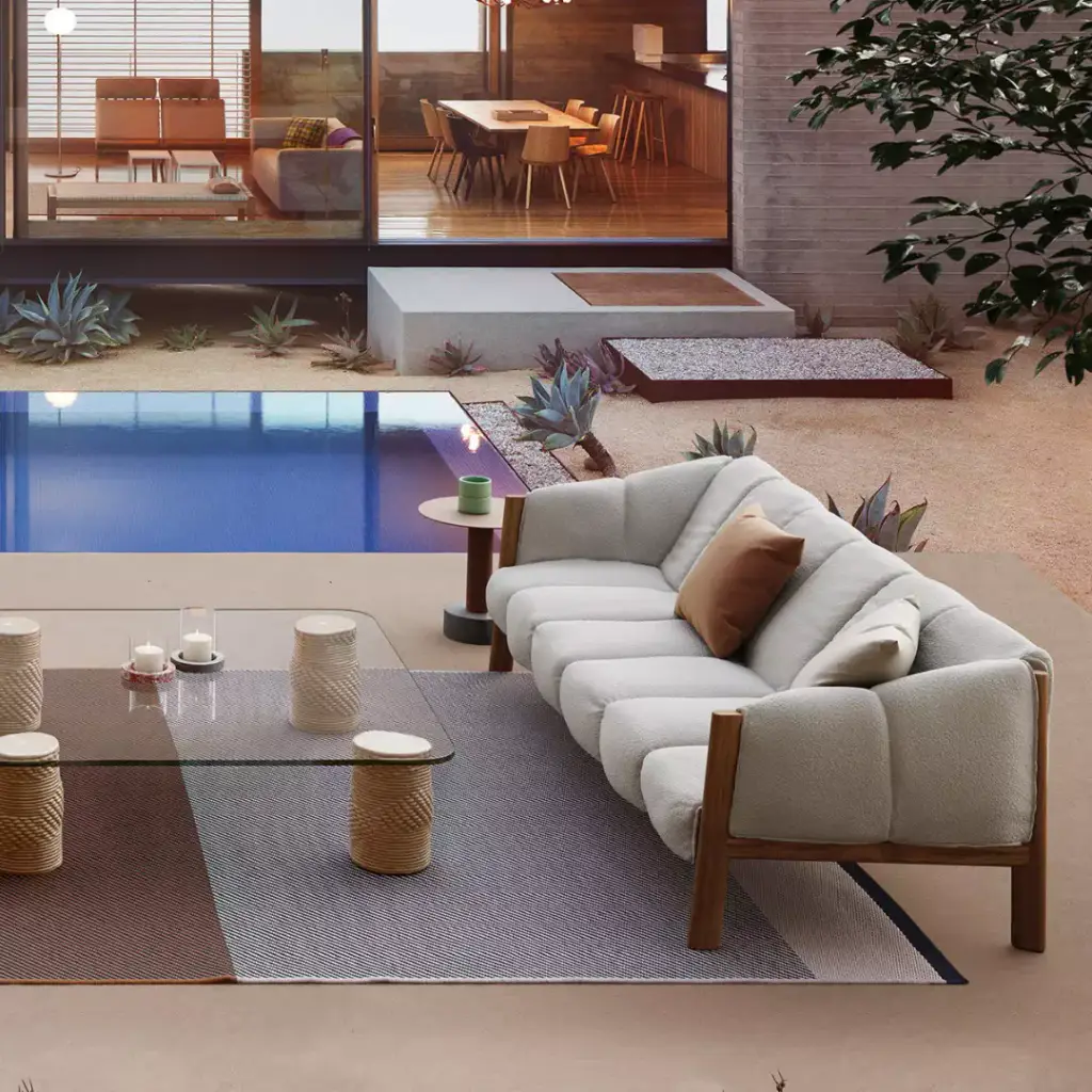 Supplier image: outdoor sofa with a wooden frame and light cushions, glass coffee table on a rug, pool and a house with lit rooms in the background.