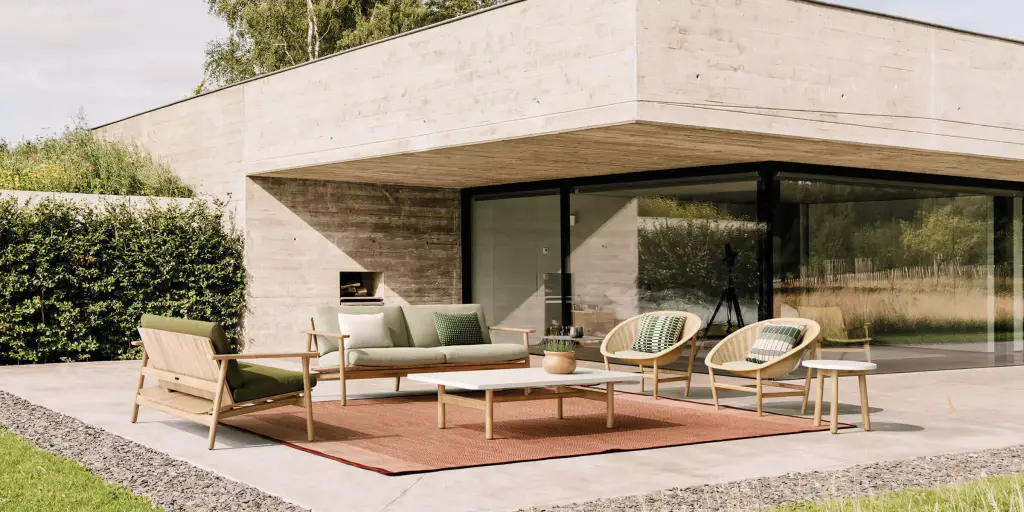 Visuel de fournisseur : salon d’extérieur bas (canapé, fauteuils cannés, table basse) posé sur un tapis rouge, devant une maison en béton brut aux grandes baies vitrées, haie taillée à gauche.