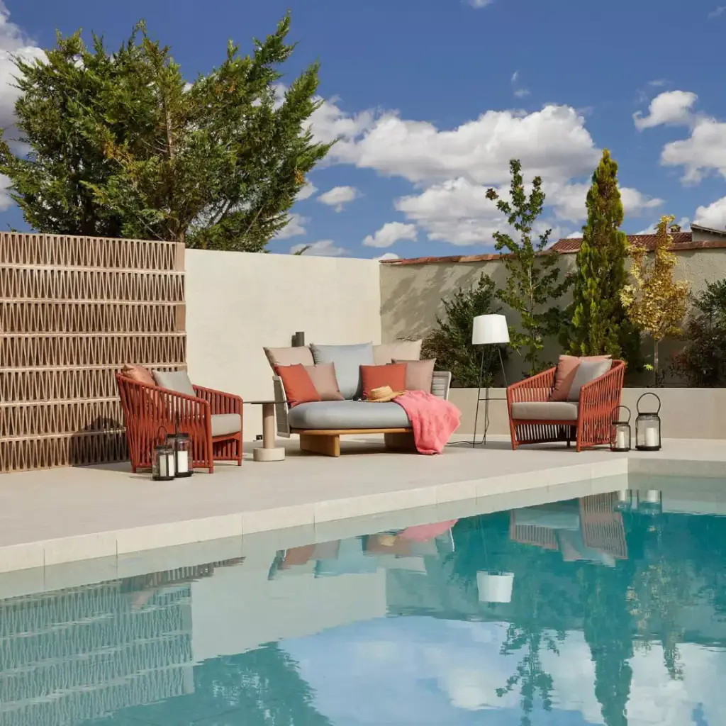 Visuel de fournisseur : salon d’extérieur au bord d’une piscine : fauteuils cordés rouges, canapé gris à coussins colorés, lampadaire, claustra en terre cuite et cyprès derrière.