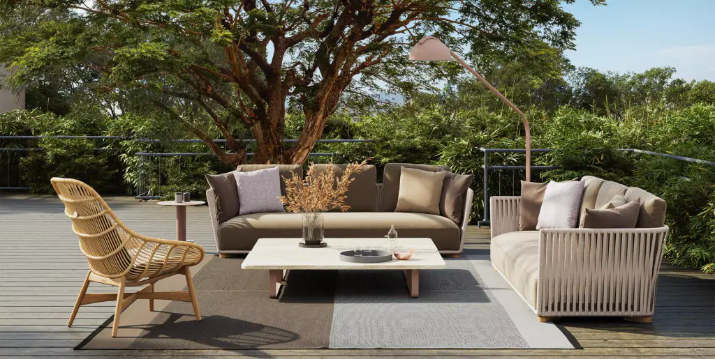 Visuel de fournisseur : salon d’extérieur sous un grand arbre : canapé et fauteuil à structure cordée, fauteuil en rotin, table basse blanche et tapis sur une terrasse en bois, lampadaire arqué.