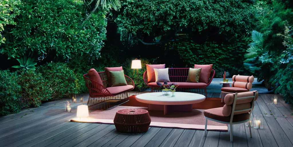 Visuel de fournisseur : salon d’extérieur à fauteuils cordés rouges et coussins colorés autour d’une table basse ronde, sur une terrasse en bois ; lanternes allumées, végétation dense à la tombée du jour.