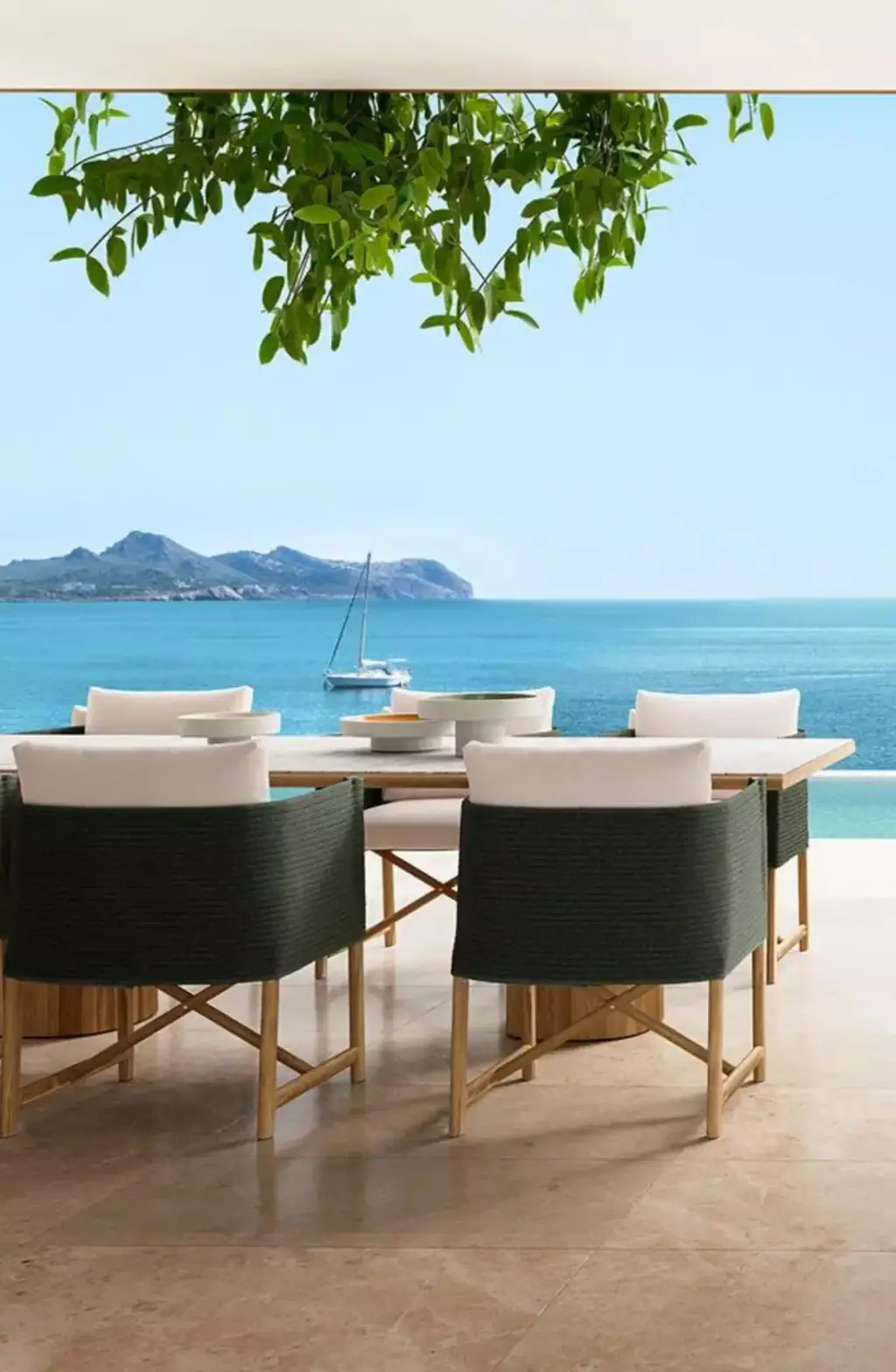 Visuel de fournisseur : table à manger d’extérieur et fauteuils vert foncé à assise claire sur une terrasse en pierre, mer turquoise, voilier et île au fond, feuillage en haut du cadre.