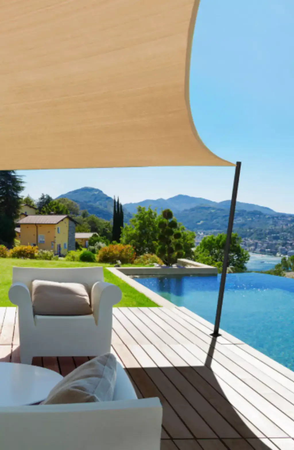 Supplier image: beige shade sail tensioned on a dark mast above a timber deck, white armchairs in the foreground, infinity pool, lake and mountains in the background.