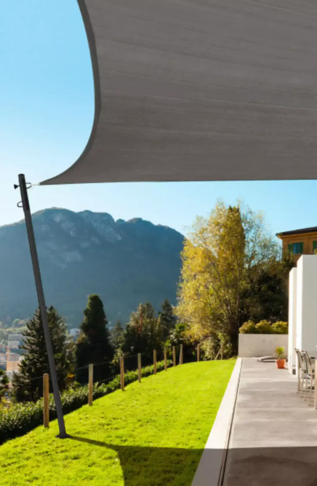 Greige shade sail stretched between the house and an angled mast, over a paved path along a sloping lawn, trees and mountain behind.