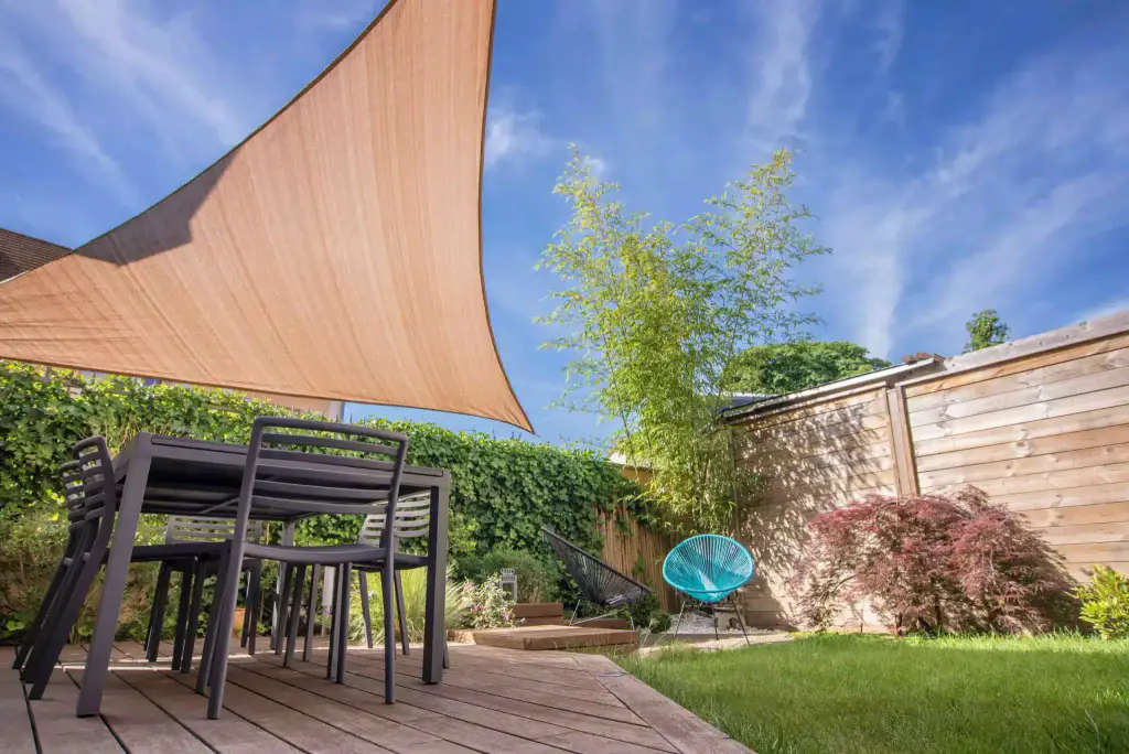 Voile d’ombrage triangulaire couleur sable tendue au-dessus d’une terrasse en bois : table et chaises anthracite, fauteuil turquoise, érable pourpre, bambou et palissade en bois, ciel bleu strié.