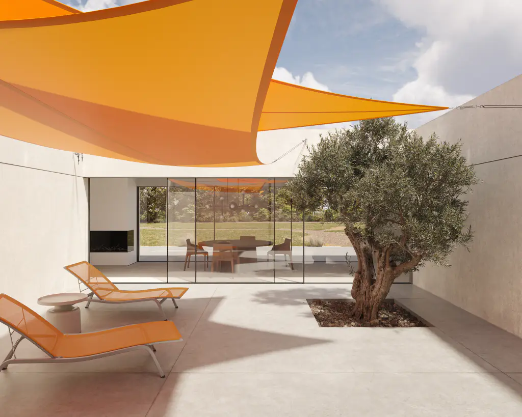 Visuel du fabricant Serge Ferrari : deux voiles d’ombrage orange tendues au-dessus d’un patio dallé, un olivier, deux bains de soleil et la baie vitrée de la maison au fond.