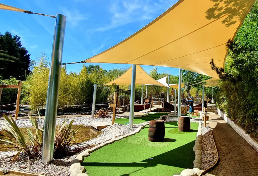 Photograph by the manufacturer Serge Ferrari: several sand-coloured triangular shade sails stretched between steel masts, over a bamboo garden under a blue sky.