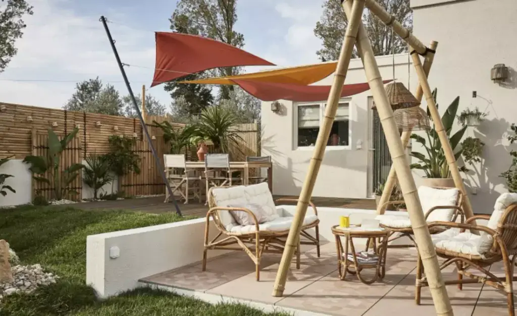 Supplier image: two red and orange sails above a rattan lounge set and a swing seat, in a garden.