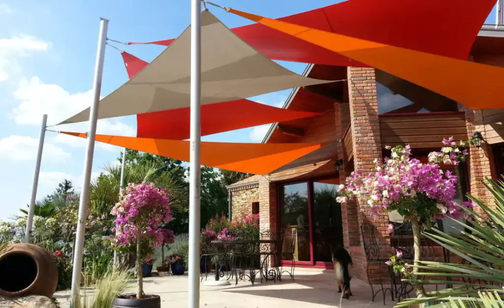 Supplier image: red, orange and greige triangular shade sails layered on white masts above a terrace, in front of a brick and timber house, bougainvillea in bloom.