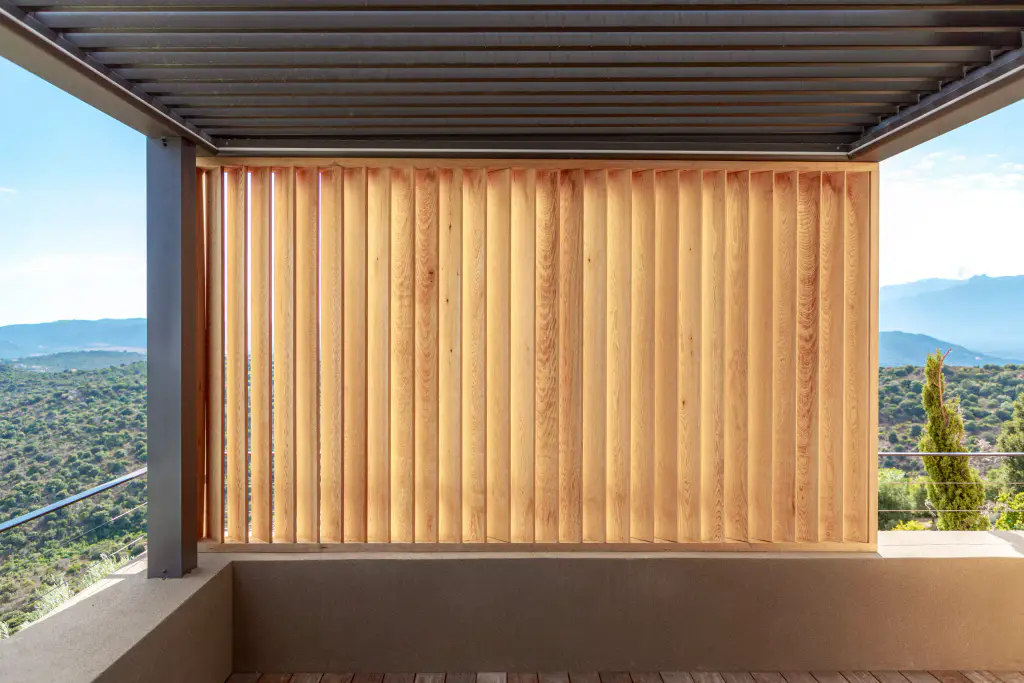 Under the anthracite pergola: a panel of vertical wooden louvres closed at the centre, maquis on either side.