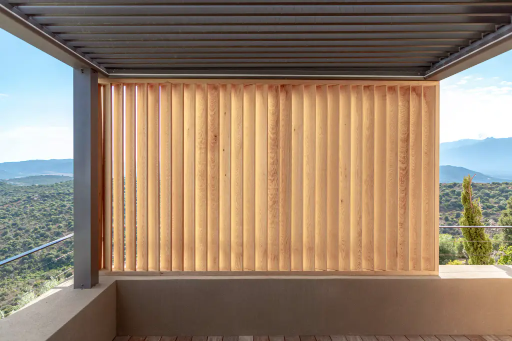 Panel of vertical wooden louvres closing the end of the terrace under the anthracite pergola, a rendered low wall in front, maquis, cypresses and mountains on either side.