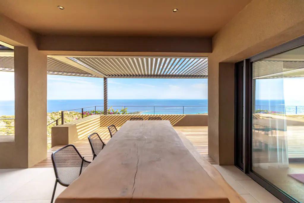 Loggia : grande table en bois massif en amorce, pergola à lames anthracite au-delà, horizon marin.