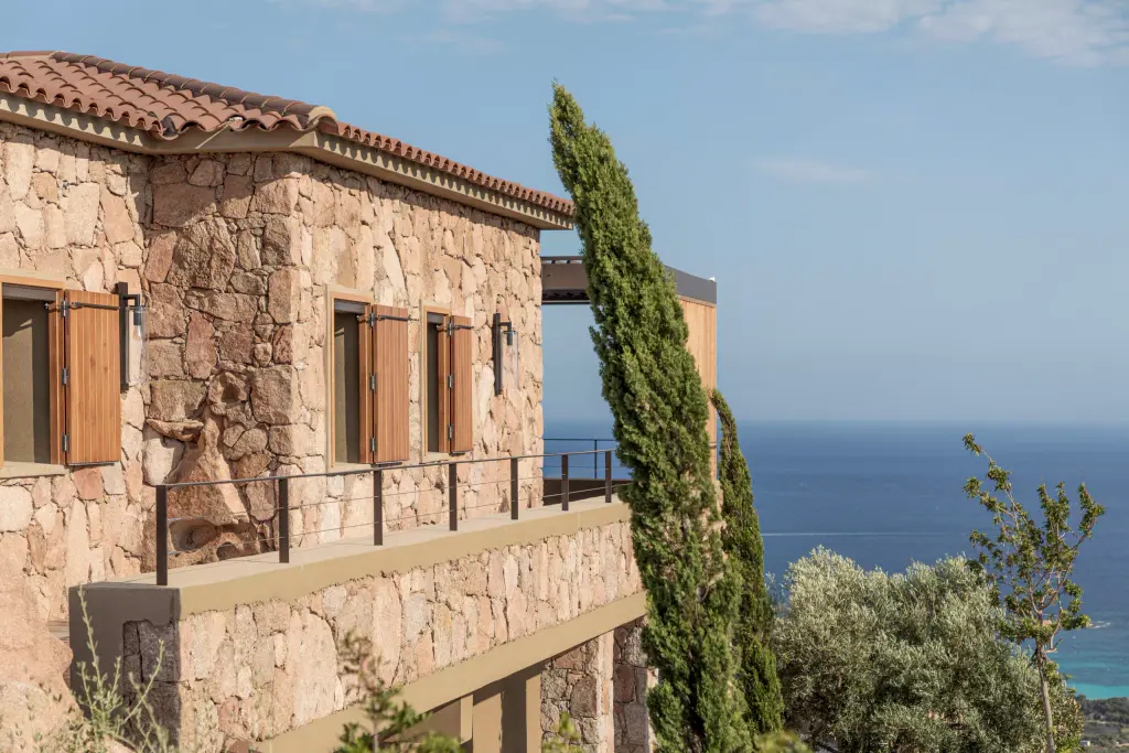 Stone house, cypresses, wooden shutters and blue sea; pergola visible at the back on the right.