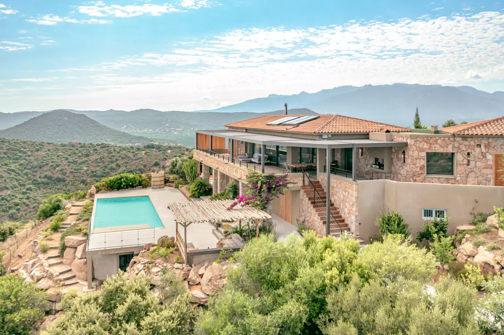 Maison en pierre perchée au-dessus du maquis, vue par drone : pergola anthracite sur la terrasse de l’étage, escalier, piscine sur son socle, rochers, collines et montagnes.
