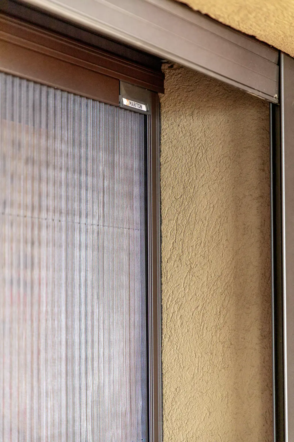 Moustiquaire plissée déployée dans l’encadrement brun d’une baie, vue de près : toile à plis fins, coulisse et étiquette de la marque en tête, mur en crépi ocre à droite.