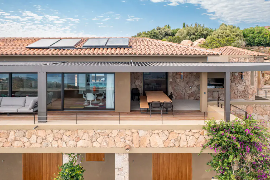 Frontal drone view: terrace under the pergola, glazed doors, wooden table, stone wall, solar panels on the roof.