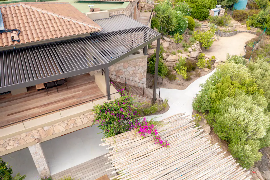 Drone view: pergola from above, tiled roof, reed screen, bougainvillea, garden.
