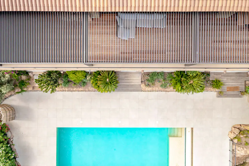 Vertical drone view: pergola louvres and deck at the top, turquoise pool at the bottom, planted beds in between.