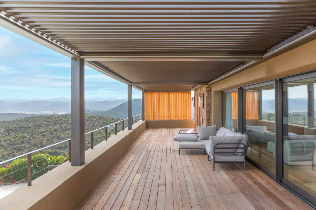 Long decked terrace under the anthracite pergola (louvres closed), grey sofa, glazed doors, wooden brise-soleil in the background, maquis on the left.