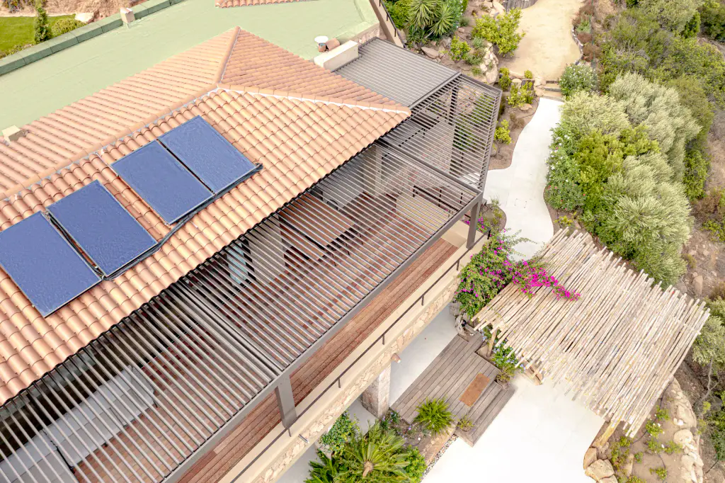 Overhead drone view: tiled roof and solar panels dominate, pergola with open louvres below.
