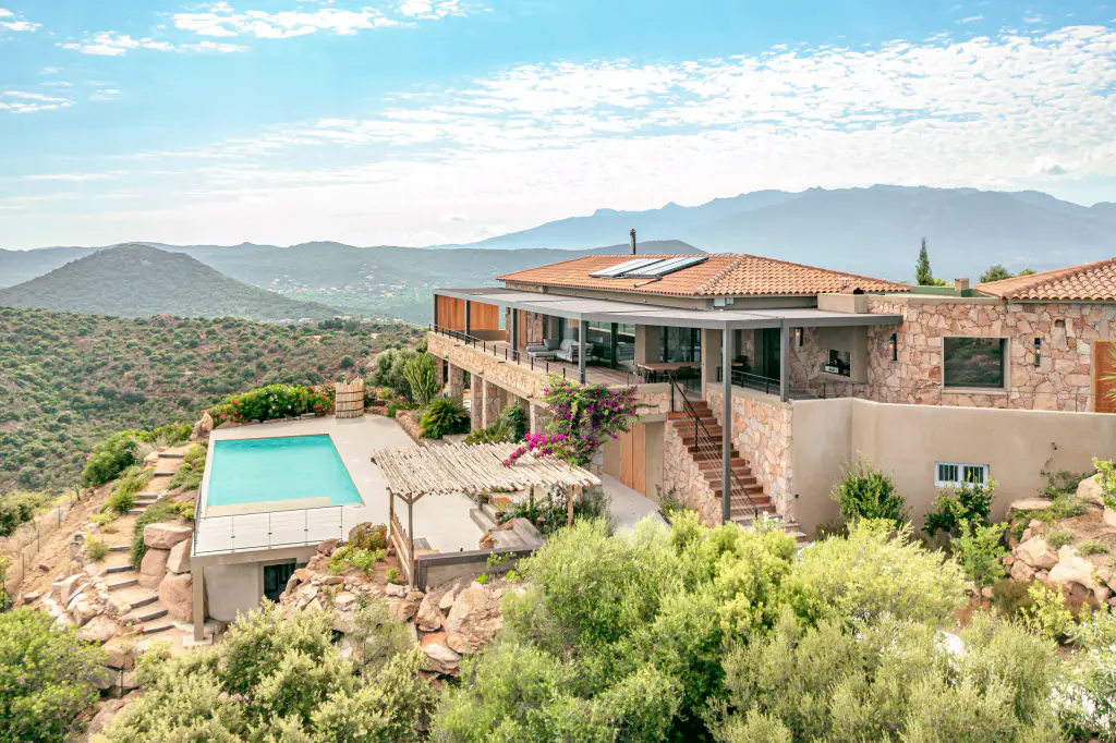 Vue drone large : villa en pierre perchée, piscine, rochers, maquis, montagnes et golfe en fond.