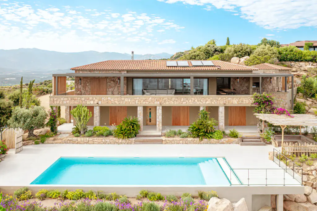 Vue drone : villa en pierre à toit de tuiles, piscine turquoise, jardin fleuri, montagnes.
