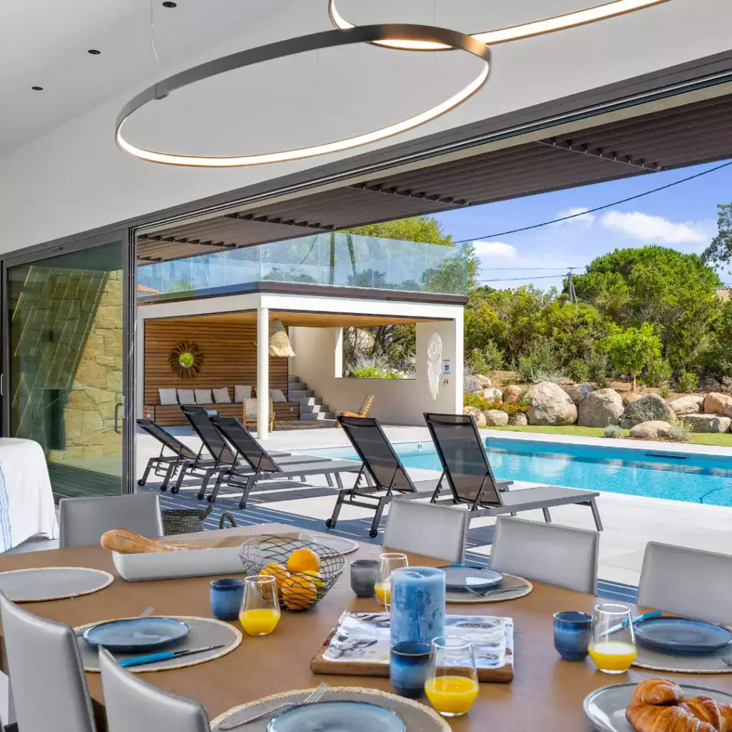 A breakfast table laid in the foreground, a circular pendant light, the glazed opening onto the pool and the sun loungers.