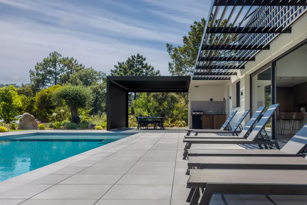 Bains de soleil gris alignés au bord de la piscine sur le dallage clair, casquette brise-soleil à droite, pergola anthracite.