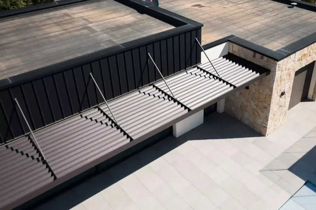 Casquette brise-soleil haubanée vue de près par drone : lames vues du dessus, tirants ancrés dans le bardage noir, mur de pierre blonde à droite, dallage gris.