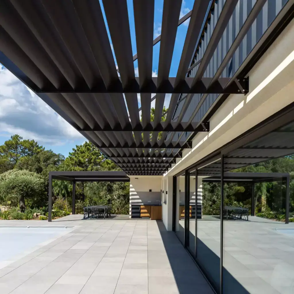 Anthracite louvred brise-soleil canopy seen from below, along the glazed façade; pale grey paving, the summer kitchen pergola in the background in front of the pines.