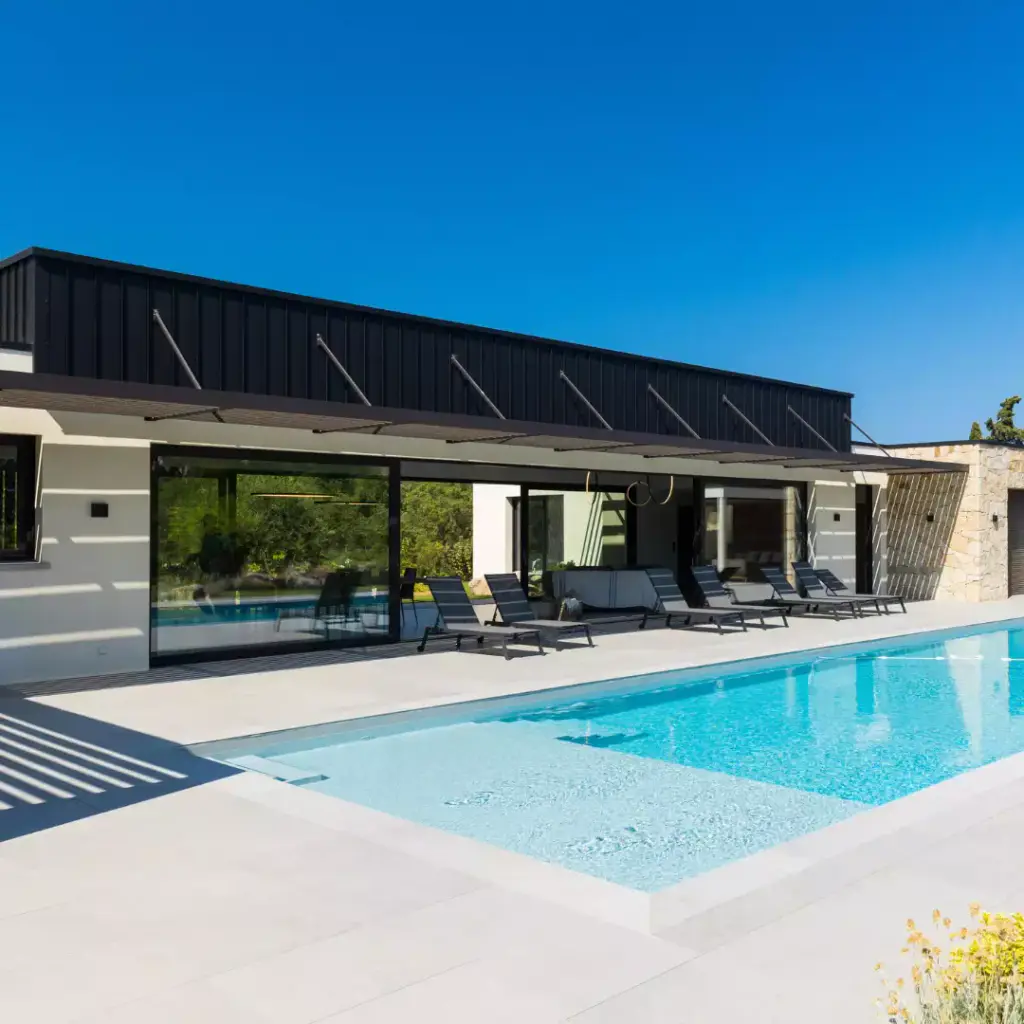 Louvred brise-soleil canopy, held by tie rods, above the glass doors of a villa clad in black zinc; sun loungers in a row and pool in the foreground, clear blue sky.