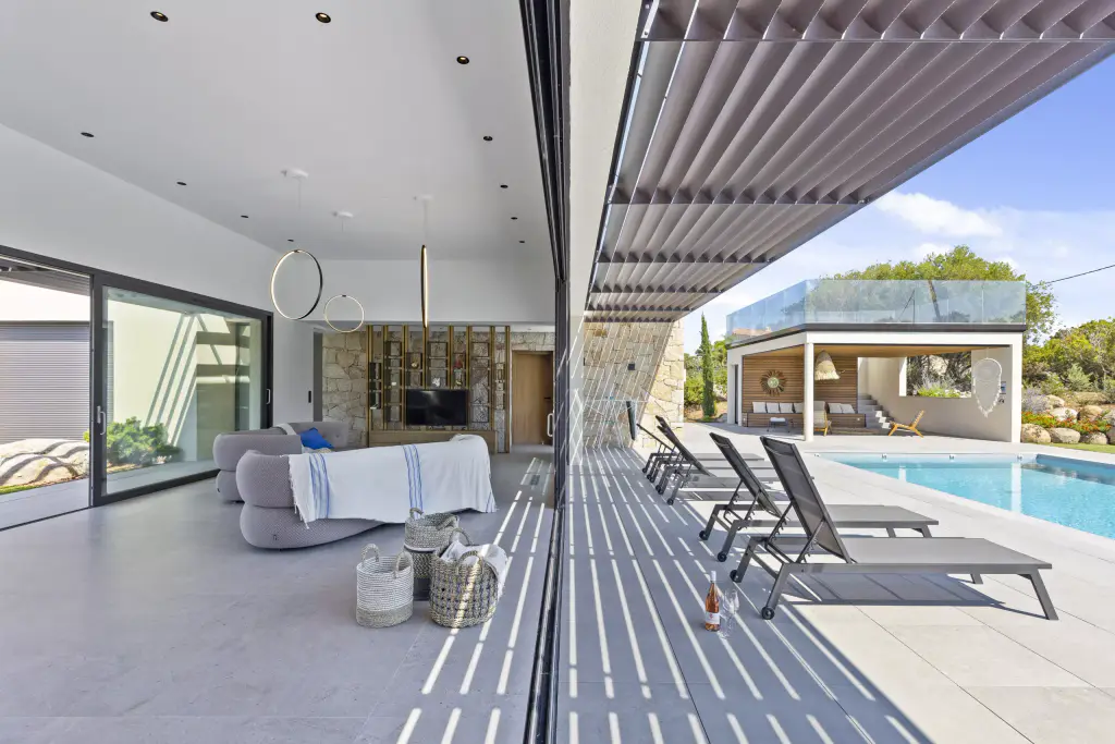 Anthracite louvred brise-soleil canopy above the wide-open glazing: its shadows stripe the paving, grey lounge on the left, sun loungers, pool and pool house on the right.