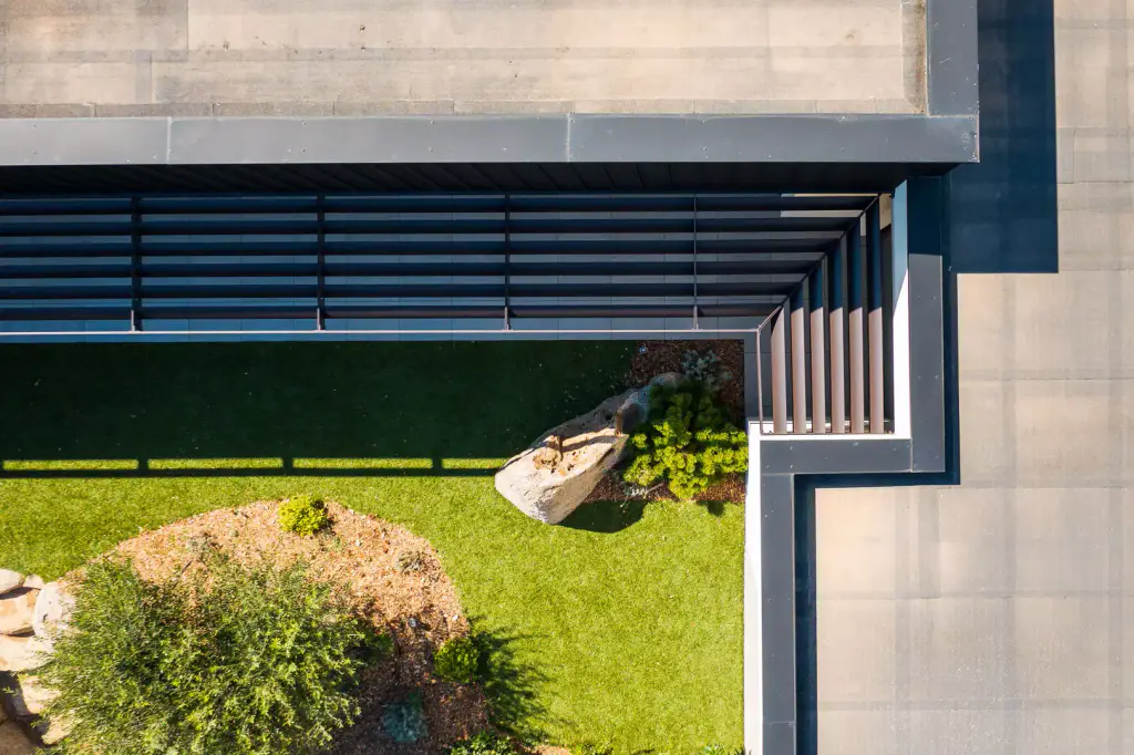 Overhead drone view: louvred brise-soleil turning the corner seen from above, lawn, rock, paving.