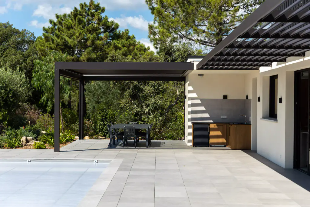 Anthracite pergola over the summer kitchen, brise-soleil canopy on the right, pines, grey paving, pool cover closed.