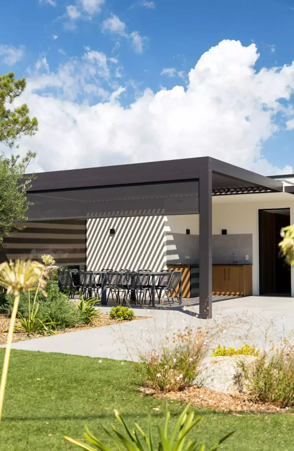Pergola anthracite abritant la cuisine d’été : un store latéral à demi descendu raye le mur d’ombres ; la table, le jardin, des cumulus.