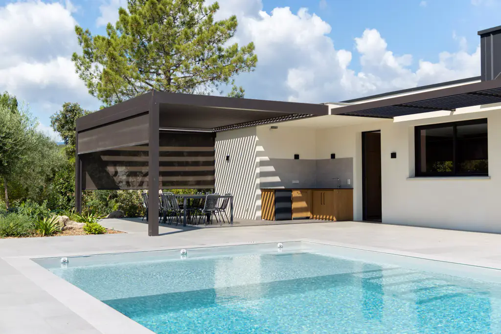 Pergola anthracite, store latéral à demi descendu, cuisine d’été, la piscine au premier plan.