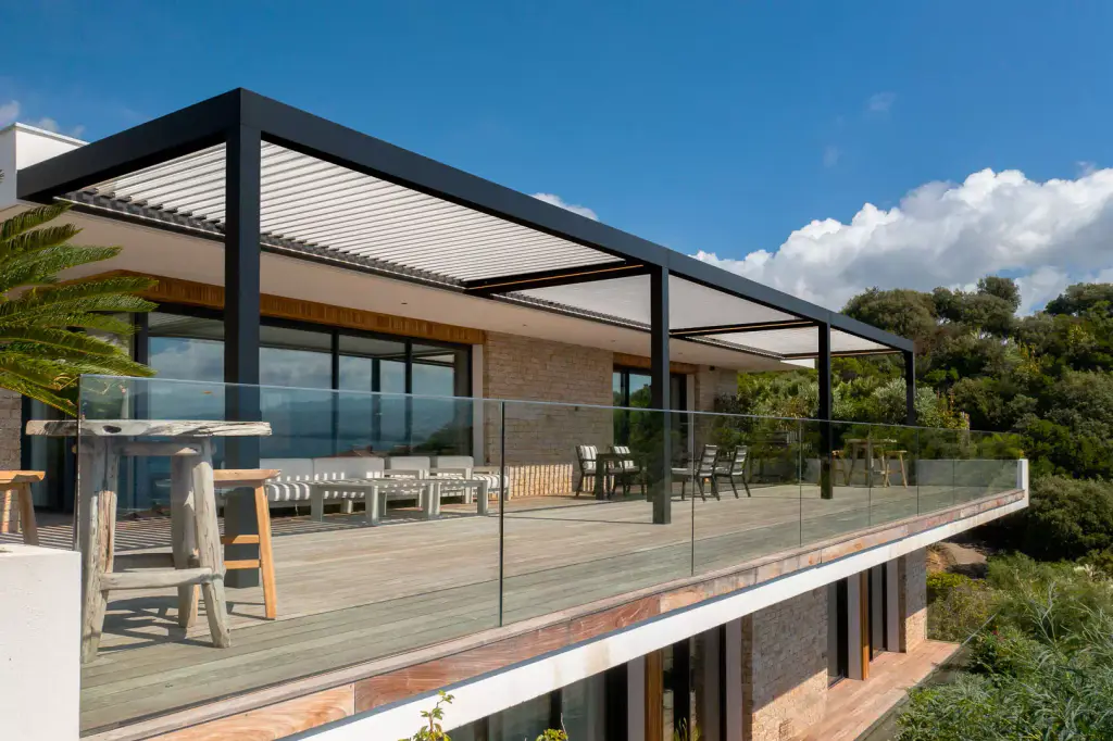Pergola bioclimatique à cadre anthracite et lames claires au-dessus d’un deck en bois : salon blanc, table et chaises, garde-corps en verre, villa en pierre, cumulus sur le maquis.
